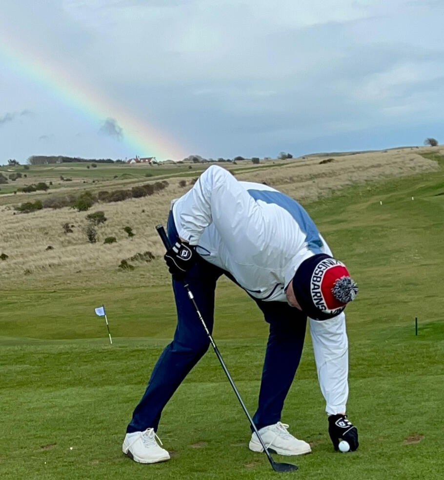 Big Wullie Golf on the course William bends to place a golf ball on a tee as a rainbow arcs across the course behind him.